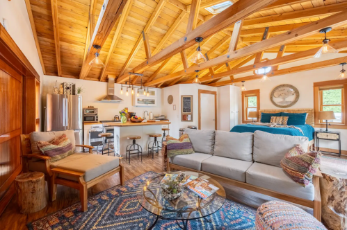 Cozy cabin interior with wooden beams, a living area, kitchen, and a bedroom, featuring warm decor and natural light.