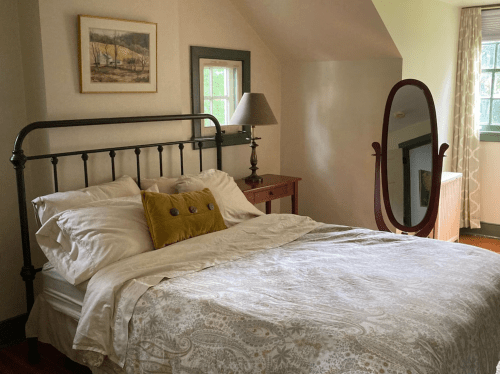 Cozy bedroom with a metal bed frame, decorative pillow, lamp, and a full-length mirror against a light-colored wall.