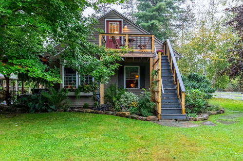 A charming two-story house with a wooden deck, surrounded by greenery and a well-maintained lawn.