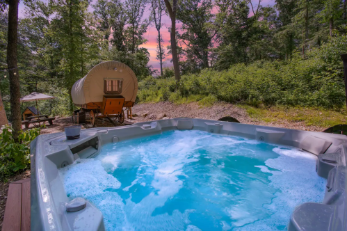 A hot tub surrounded by trees, with a cozy wagon in the background and a colorful sunset sky.