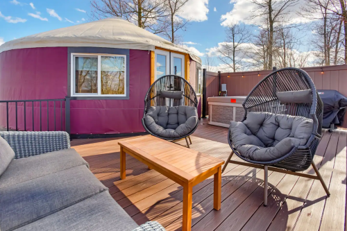 A cozy outdoor deck with a yurt, two black chairs, a wooden table, and a hot tub in a scenic setting.