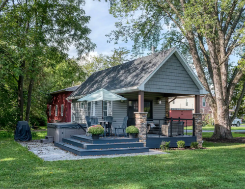 A cozy gray house with a patio, surrounded by trees and a red building in the background, on a sunny day.