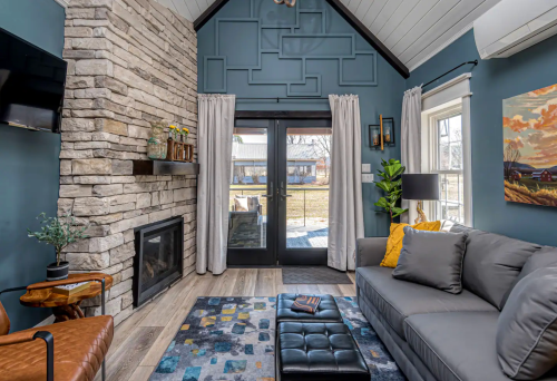Cozy living room with stone fireplace, blue walls, large windows, and modern furniture, featuring a view of the outdoors.