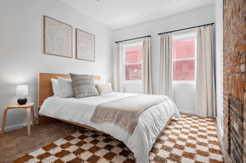 Cozy bedroom with a bed, decorative pillows, a lamp, and large windows, featuring a textured rug and exposed brick wall.