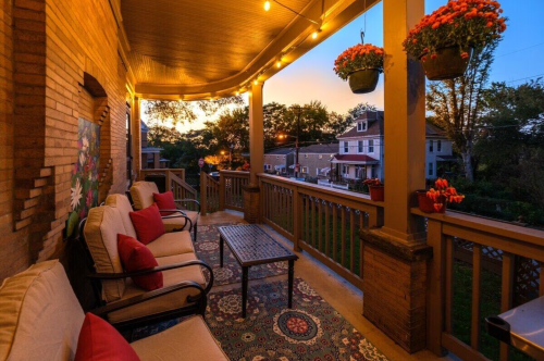 Cozy porch with seating, potted flowers, and warm lights, overlooking a neighborhood at sunset.