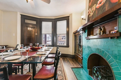 A dining room with a glass table set for dinner, featuring a green tiled fireplace and large windows.