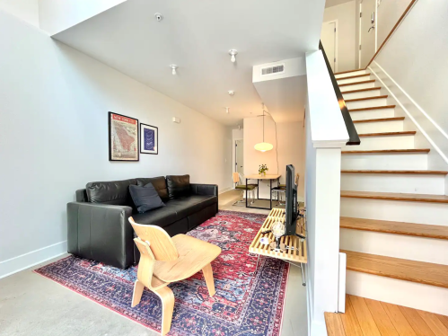 Modern living room with a black sofa, wooden chair, rug, and stairs leading to an upper level. Bright and airy space.