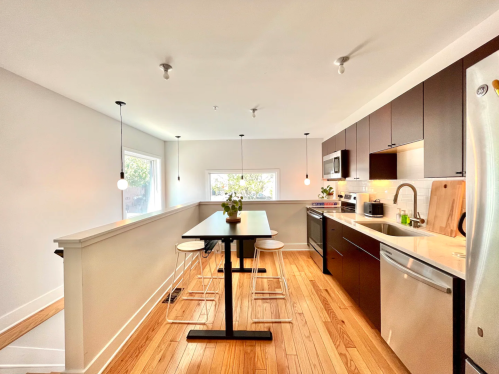Modern kitchen with dark cabinets, a light wood floor, a dining table, and large windows letting in natural light.