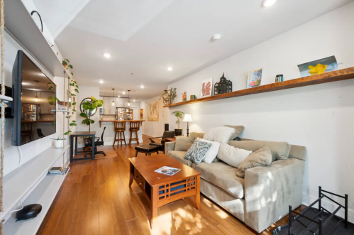 A cozy living room with a sofa, coffee table, and shelves, featuring plants and a TV in a bright, modern space.