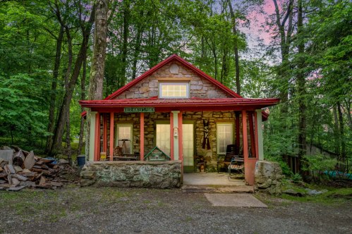 A charming stone cabin with a red roof, surrounded by lush green trees and a gravel path.