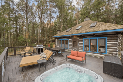 Cozy log cabin with a deck, hot tub, outdoor seating, and grill, surrounded by trees in a serene setting.