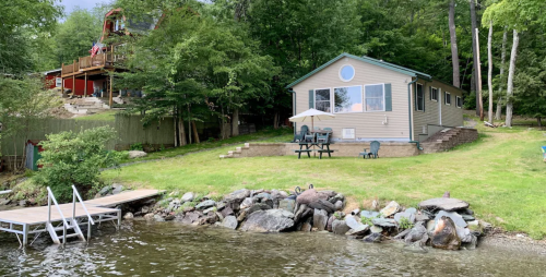 A lakeside cottage with a patio, surrounded by greenery and a dock by the water.