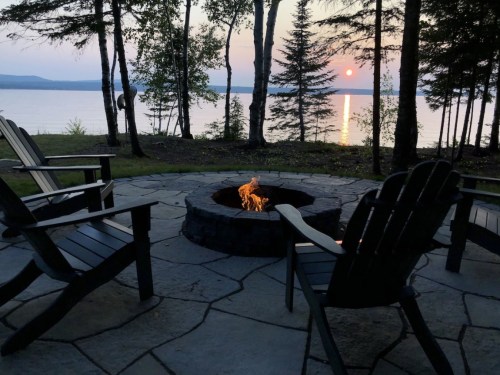 A serene lakeside scene at sunset, featuring a fire pit surrounded by wooden chairs and trees.