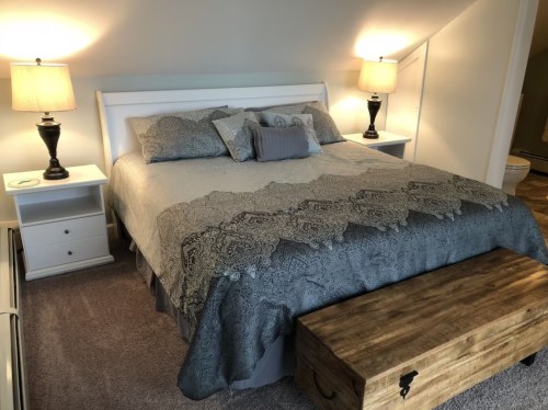 Cozy bedroom featuring a queen bed with a patterned gray blanket, two lamps, and a wooden storage bench at the foot.