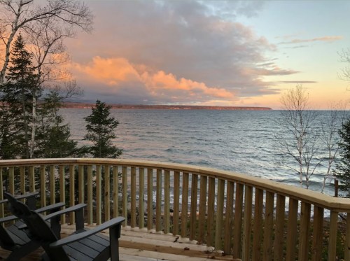 Scenic view of a lake at sunset, with trees and a wooden deck in the foreground. Soft clouds reflect warm colors.