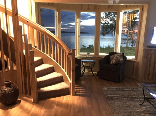 Cozy interior with a spiral staircase, armchair, and large windows overlooking a serene lake at sunset.