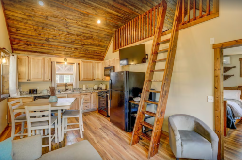 Cozy cabin interior featuring a kitchen, dining area, and a wooden staircase leading to a loft. Warm, rustic decor.