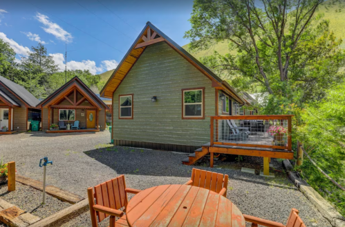 A cozy cabin with a wooden deck, surrounded by trees and a gravel area, under a bright blue sky.