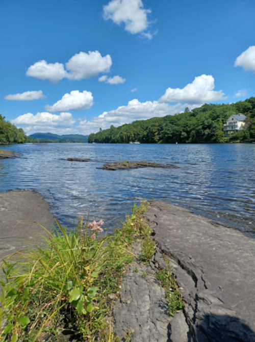 A serene lake scene with rocky shores, lush greenery, and fluffy clouds under a bright blue sky.