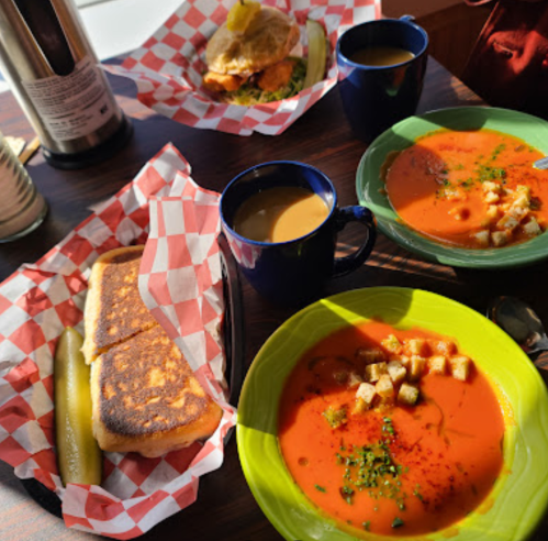 A table with two bowls of tomato soup topped with croutons, a sandwich, coffee, and a pickle on a checkered paper.