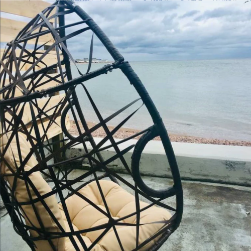 A cozy hanging chair with a woven design overlooks a calm sea under a cloudy sky.