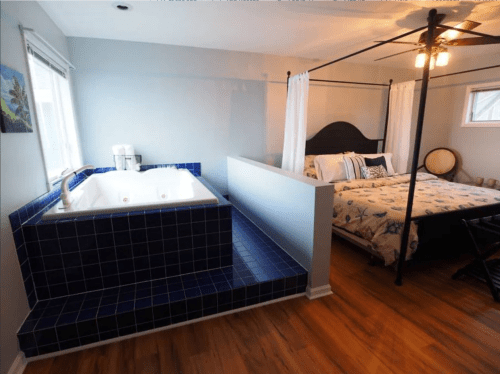 A cozy bedroom featuring a four-poster bed and a blue-tiled jacuzzi area. Natural light fills the space.