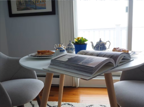 A cozy table setting with a book, pastries, a teapot, and a potted plant by a window with natural light.