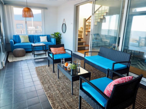 Bright sunroom with blue furniture, a woven rug, and large windows, featuring a staircase in the background.