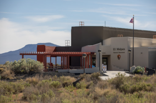 El Malpais National Monument visitor center with a mountain backdrop and American flag flying. Desert vegetation in foreground.
