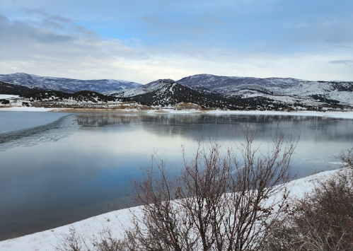 A serene winter landscape featuring a calm lake, snow-covered mountains, and a cloudy sky.