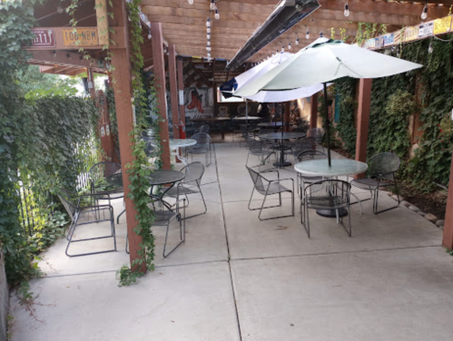 Outdoor seating area with metal tables and chairs, shaded by umbrellas, surrounded by greenery and string lights.