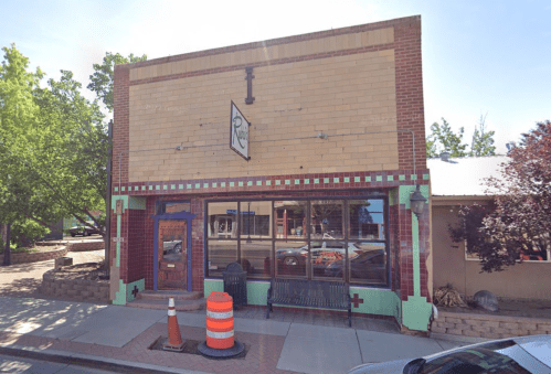 A brick building with a colorful facade, large windows, and a bench outside, set in a tree-lined street.
