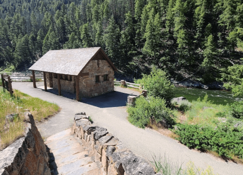A rustic stone building beside a river, surrounded by lush green trees and a gravel path leading down to the water.