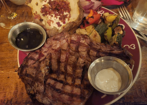 A plate featuring a grilled steak, baked potato with toppings, and a side of mixed grilled vegetables.