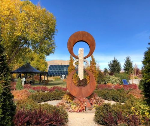 A modern sculpture surrounded by colorful autumn foliage and greenery under a clear blue sky.