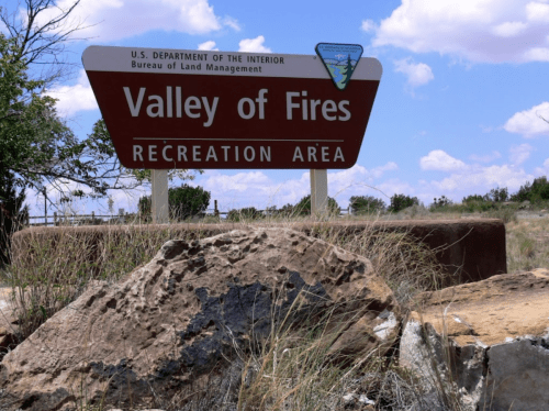 Sign for Valley of Fires Recreation Area, with blue sky and scattered clouds in the background. Rocks in the foreground.