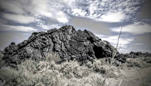 A large, textured volcanic rock formation under a cloudy sky, surrounded by sparse vegetation.