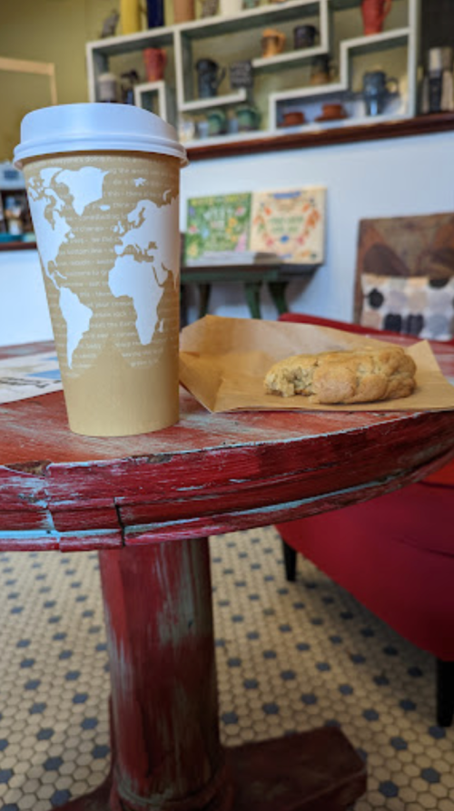 A coffee cup with a world map design sits on a rustic table next to a cookie in a cozy café setting.