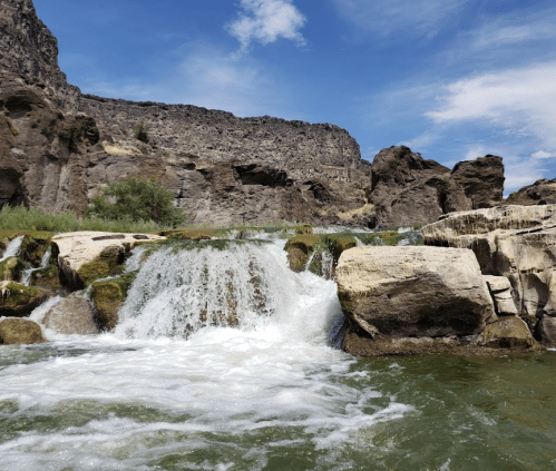 A serene waterfall cascades over rocks, surrounded by lush greenery and rugged cliffs under a blue sky.