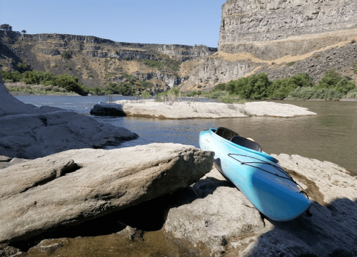 A blue kayak rests on rocky shore by a calm river, surrounded by cliffs and greenery under a clear blue sky.
