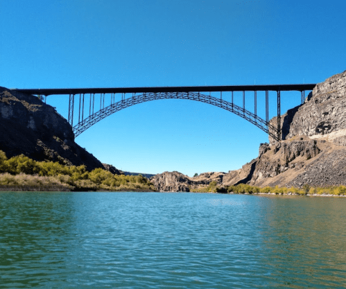 A large arch bridge spans over a calm river, surrounded by rocky cliffs and clear blue skies.