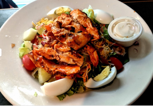 A colorful salad topped with grilled chicken, cheese, tomatoes, eggs, and a side of dressing.