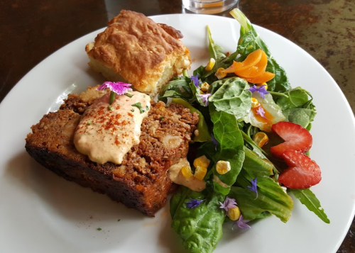 A plate with meatloaf topped with sauce, a piece of cornbread, and a fresh salad with flowers and strawberries.