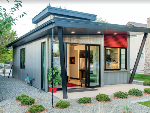 Modern metal-clad house with large windows, a red accent wall, and landscaped gravel yard.