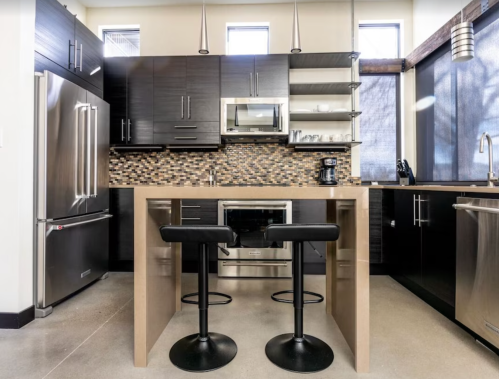 Modern kitchen with dark cabinetry, stainless steel appliances, a mosaic backsplash, and two black bar stools at a counter.