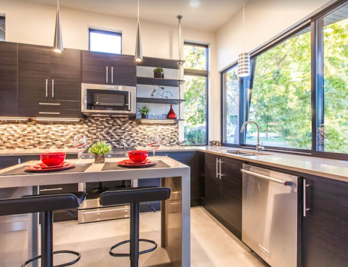 Modern kitchen with dark cabinetry, stainless steel appliances, and large windows overlooking greenery. Dining area set for two.