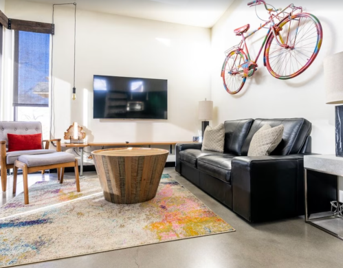 A modern living room with a black sofa, wooden coffee table, colorful rug, and a bicycle art piece on the wall.