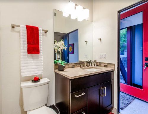 Modern bathroom featuring a sleek vanity, mirror, red accents, and a bright entrance door.