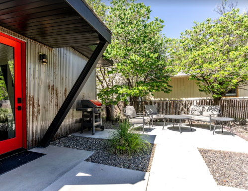 A modern outdoor patio with a grill, seating area, and lush greenery surrounding a stylish home entrance.