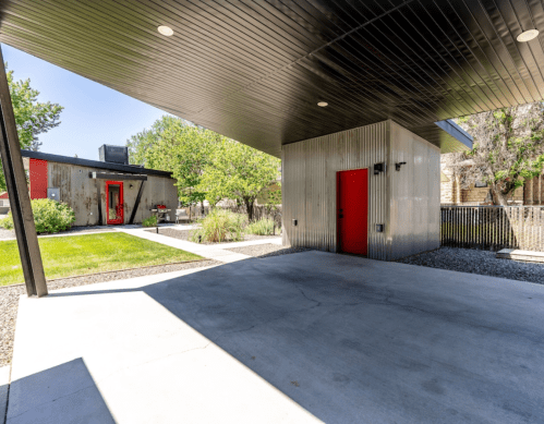 Modern outdoor space featuring a sleek metal structure, vibrant red doors, and landscaped greenery under a shaded roof.